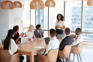 Affärsmöte i modernt konferensrum; ledare presenterar för team runt träbord med laptops och stora panoramafönster.