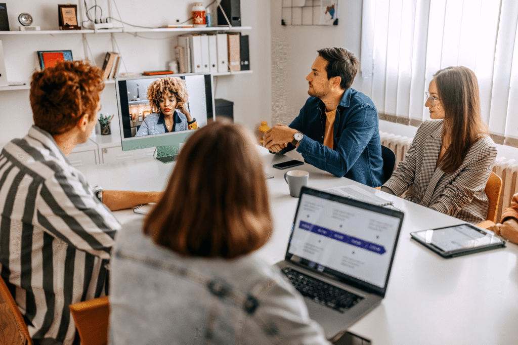 Team i hybridmöte på kontor – grupp runt mötesbord i videokonferens med kollega på skärm; laptops, surfplatta och anteckningar.