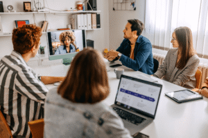 Team i hybridmöte på kontor – grupp runt mötesbord i videokonferens med kollega på skärm; laptops, surfplatta och anteckningar.