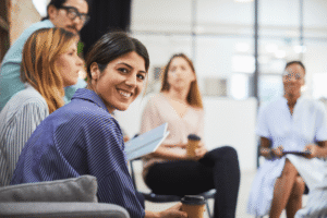 Leende kvinna i fokus under informellt teammöte; kollegor med kaffekoppar i modern kontorsmiljö – workshop och samarbete.